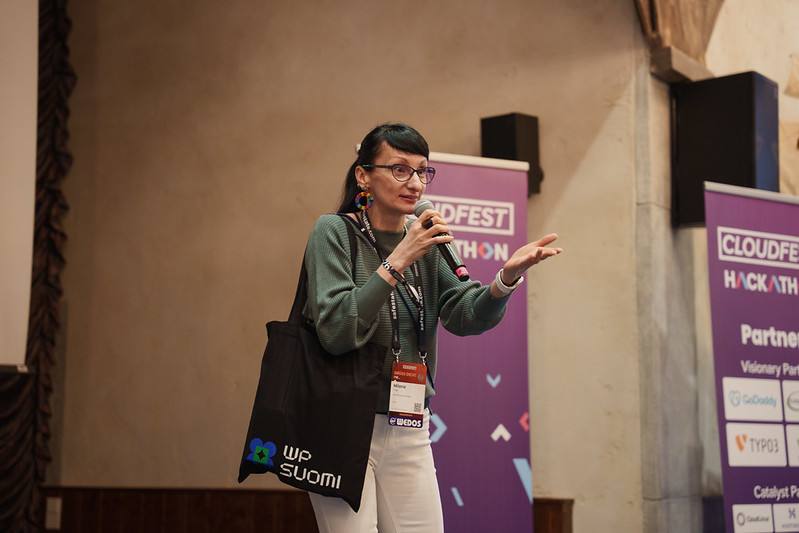 Milana Cap presenting the project pitch at CloudFest Hackathon stage, holding the WP Suomi tote bag, full of cookies.